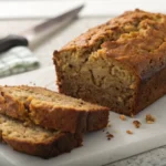 A whole loaf of delicious banana bread with olive oil on a board, with a slice cut and key ingredients in the background.