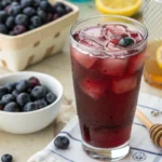 Refreshing blueberry lemonade punch in a tall glass with ice and a lemon slice, surrounded by fresh blueberries.