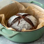 A beautiful round loaf of chocolate sourdough bread, freshly baked and dusted with flour, in a teal Dutch oven.
